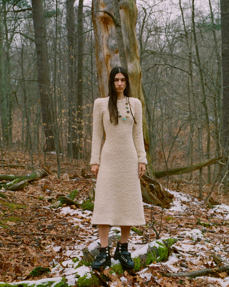 A woman in a cream-colored wool dress stands in a snowy, leaf-covered forest. She wears black boots and a necklace, and behind her are tall trees with bare branches. The atmosphere is serene and wintery.