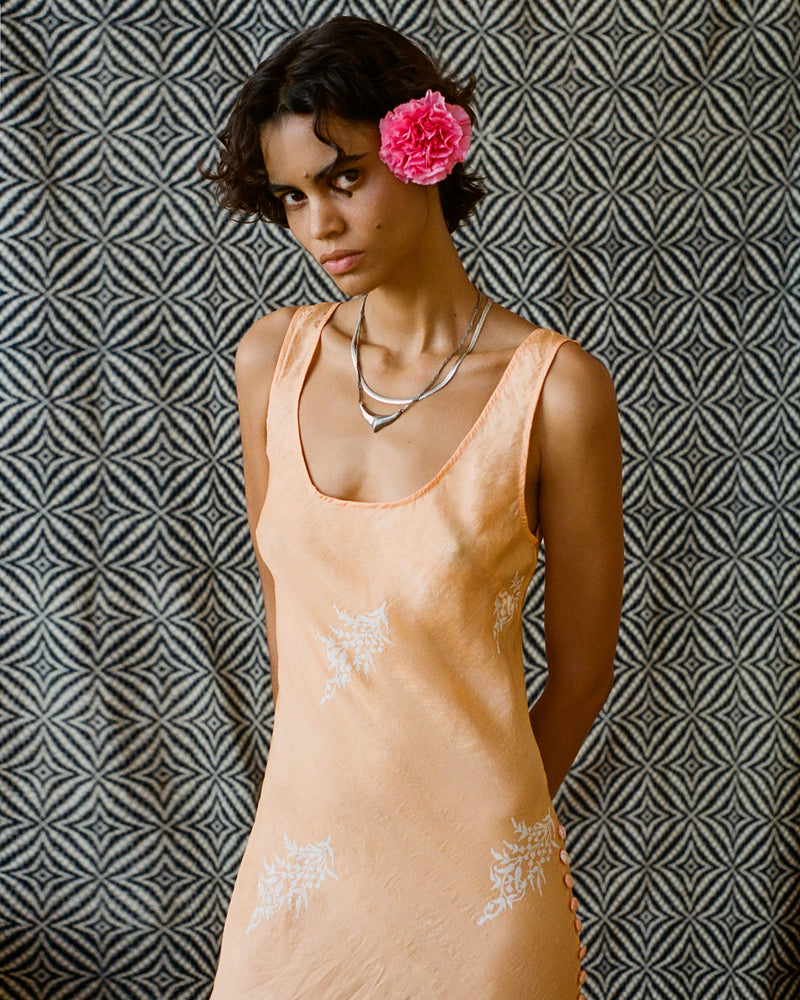 A person wearing a peach-colored dress with white embroidery stands against a black and white geometric patterned background. They have a pink flower tucked in their hair and are wearing layered necklaces.