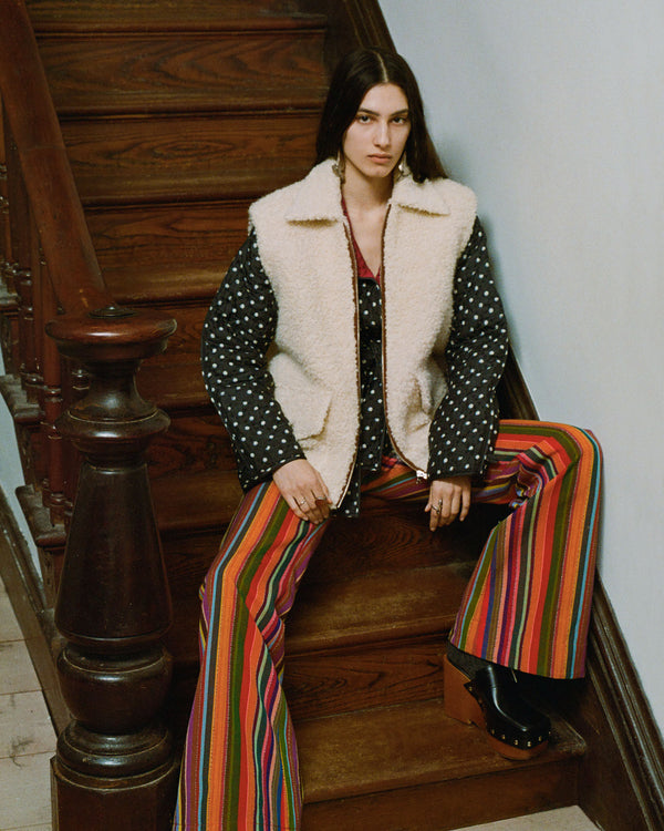 A person sits on wooden stairs wearing a polka dot jacket with a shearling collar over a red top, and colorful striped pants with platform shoes. The setting is indoors with a vintage feel.