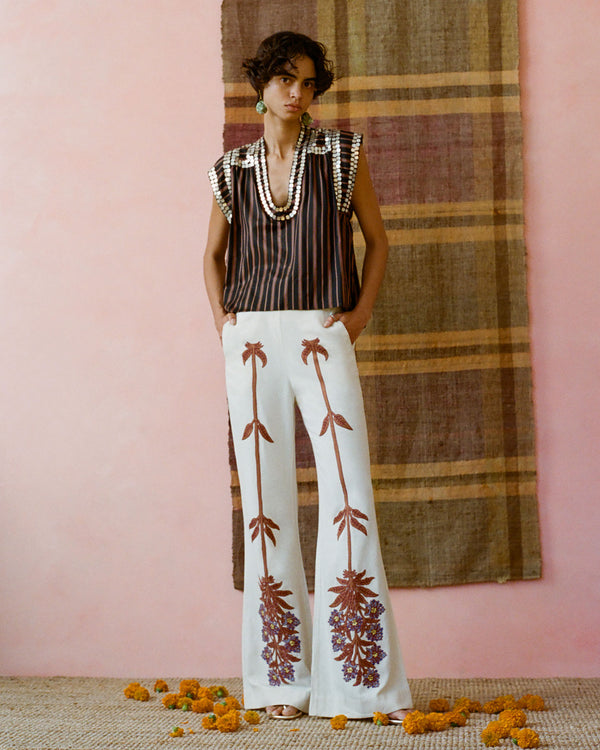 A person stands in front of a pink wall with a striped cloth backdrop. They wear a striped top with pearl details and white pants with floral designs. Orange flowers are scattered on the floor.