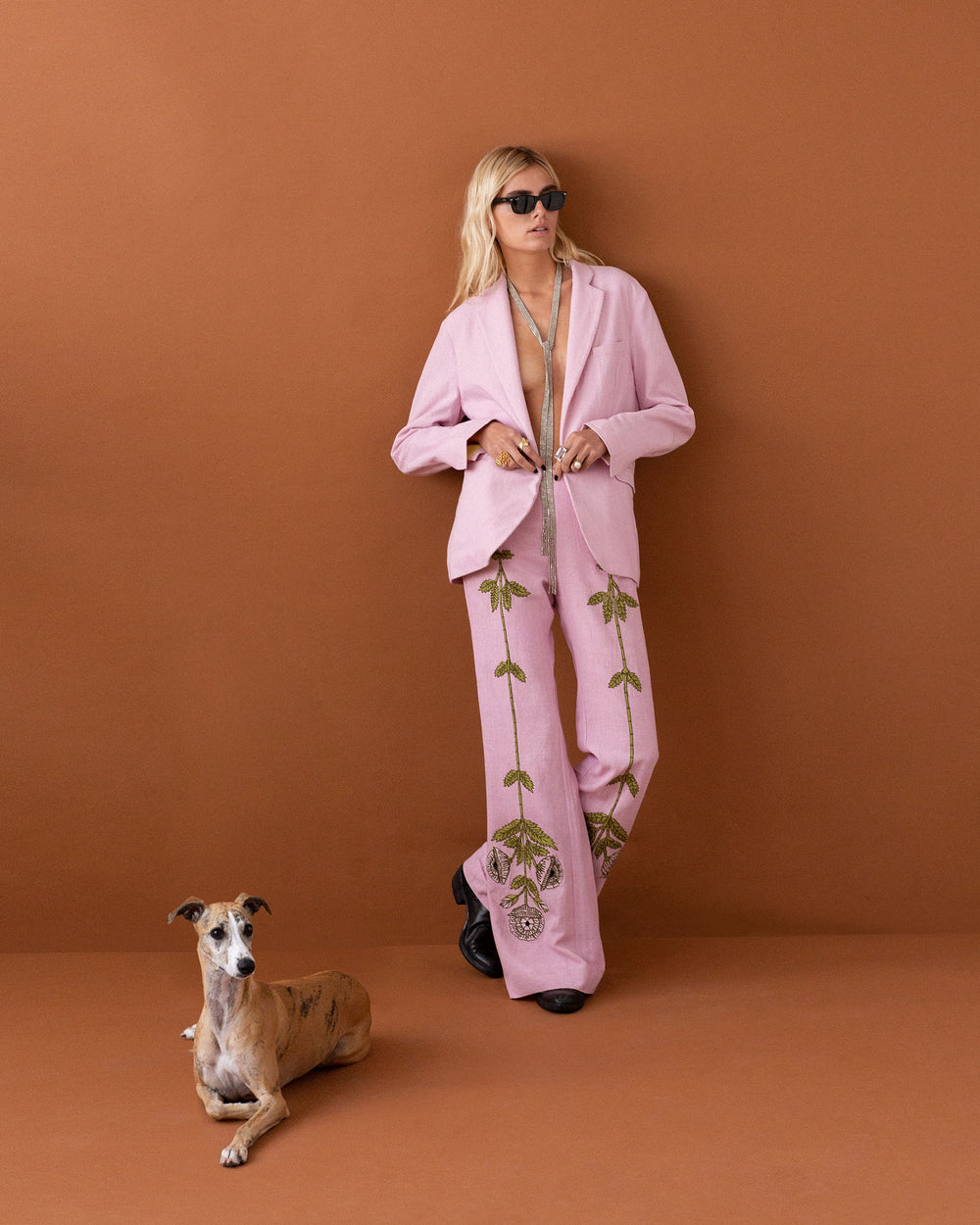 A person in a Charlie Crocus Embroidered Pant with a floral motif poses against a brown backdrop, wearing sunglasses. A short-haired dog sits nearby, gazing toward the camera.