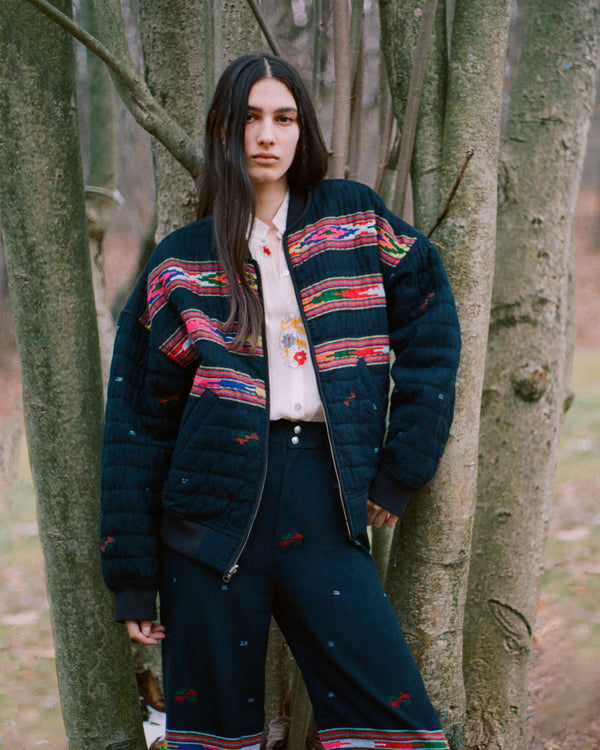 A person with long hair stands against a backdrop of tree trunks. They are wearing a dark embroidered jacket and pants with colorful patterns, and a light-colored shirt. The setting has a natural, outdoor atmosphere.