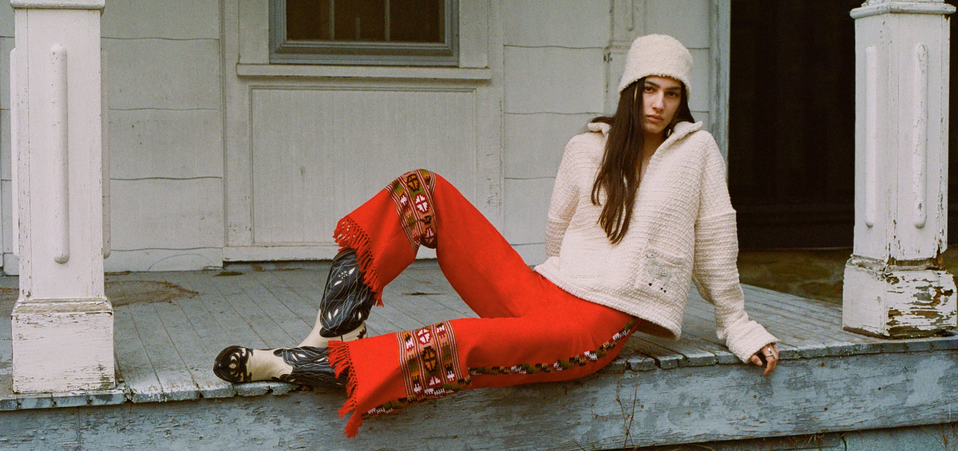 A person with long hair, wearing a knit hat, a cream sweater, and red fringed pants, sits on a wooden porch. The porch features white pillars and walls, and the person has a relaxed posture.