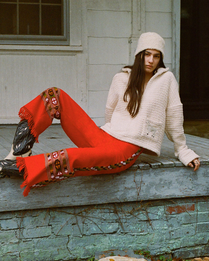 A person in a cream sweater and hat with long dark hair sits on wooden steps. They wear bright red pants with embroidered patterns and black shoes. The background is a rustic wall with a window.