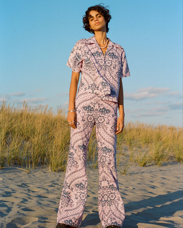 A person with short curly hair stands on a sandy beach wearing a matching pastel purple outfit with intricate patterns. In the background, tall grasses sway under a clear blue sky with a few clouds.