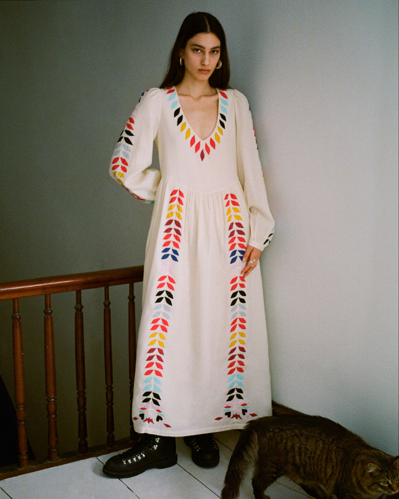 A woman stands in a white dress with colorful geometric patterns near a staircase. She has long brown hair and is wearing black shoes. A brown cat is at her feet. The background is a plain wall.