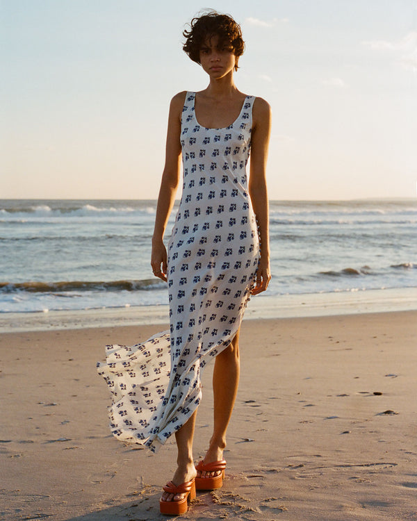 A person wearing a long, sleeveless dress with a blue pattern walks along a sandy beach. The dress flows in the breeze, and they are wearing platform sandals. The ocean and sky create a serene backdrop in the warm lighting.