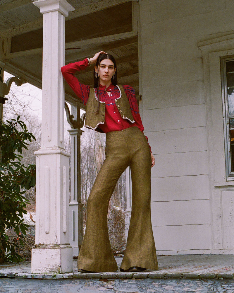 A person stands on a porch in front of a wooden house, wearing a red shirt and brown vest with matching high-waisted wide-leg pants. They are leaning against a column, with one hand resting on their head, exuding a vintage, stylish vibe.