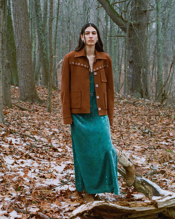 A person with long hair stands in a forest, wearing a brown jacket and a long turquoise dress. They are surrounded by trees and fallen leaves, with patches of snow on the ground.