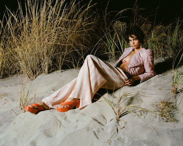 A person lounges on sand surrounded by tall grass at night. They wear a pink outfit with wide-legged pants and red platform sandals, exuding a relaxed and stylish vibe. The scene has a serene, natural backdrop.