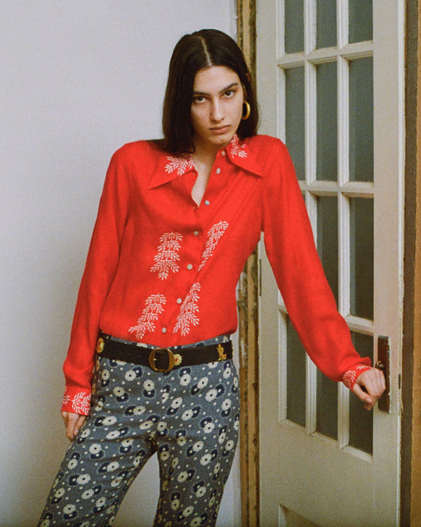 A person with shoulder-length dark hair leans against a door, wearing a red button-up shirt with white embroidery and patterned pants. They accessorize with hoop earrings and a belt. The expression is neutral or relaxed.