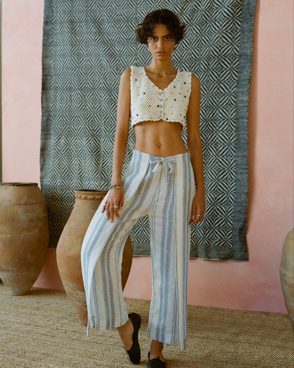 A person with short curly hair wears a white crochet crop top and blue-striped wide-leg pants. They stand on a woven rug in front of large clay pots and a patterned textile hanging on a pink wall.