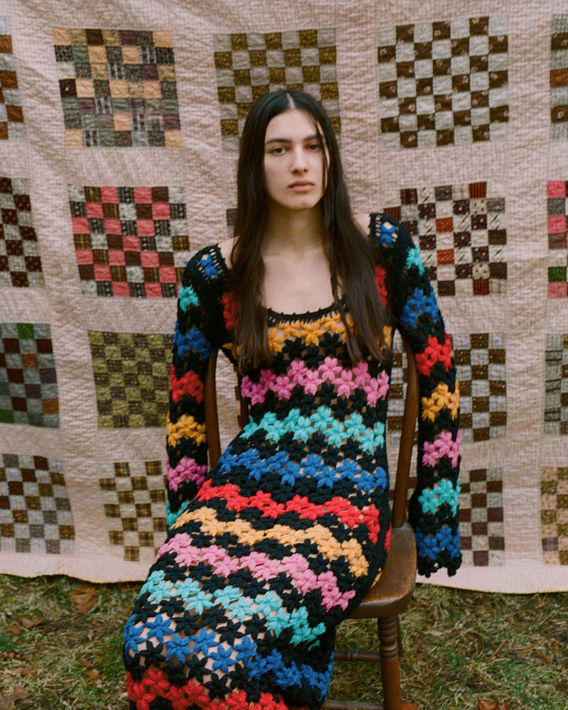 A person with long dark hair sits on a wooden chair, wearing a colorful crocheted dress. The background features a quilt with checkerboard patterns. The setting is outdoors on grass.