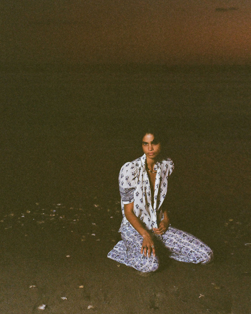 A person kneels on a beach at dusk, dressed in a patterned outfit with a white top and blue pants. The dark sky contrasts with the illuminated figure, capturing a serene and contemplative mood.