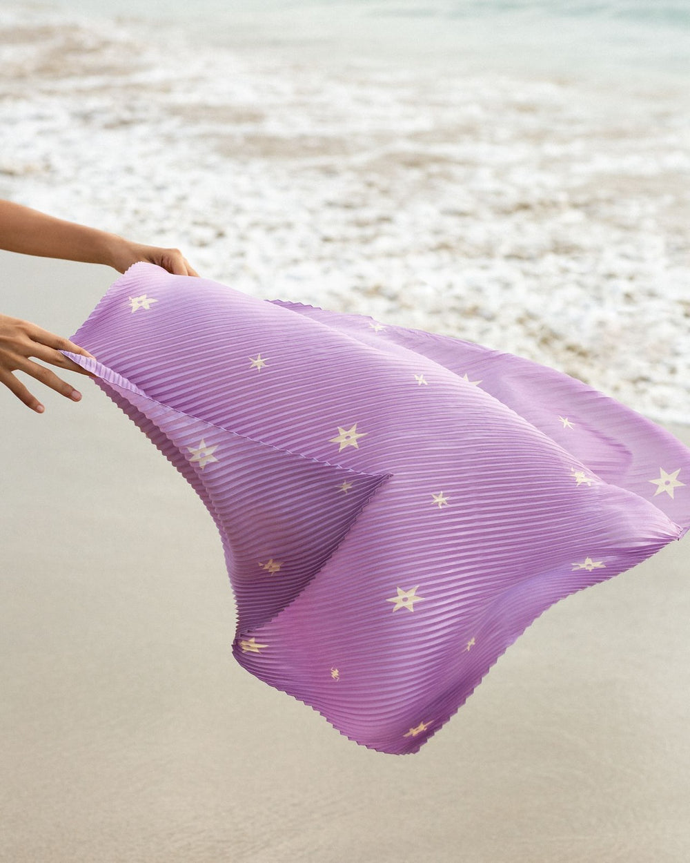 A person holds the Estella Thistle Plisse Scarf, adorned with star patterns, against a sandy beach and gentle ocean waves in the background.