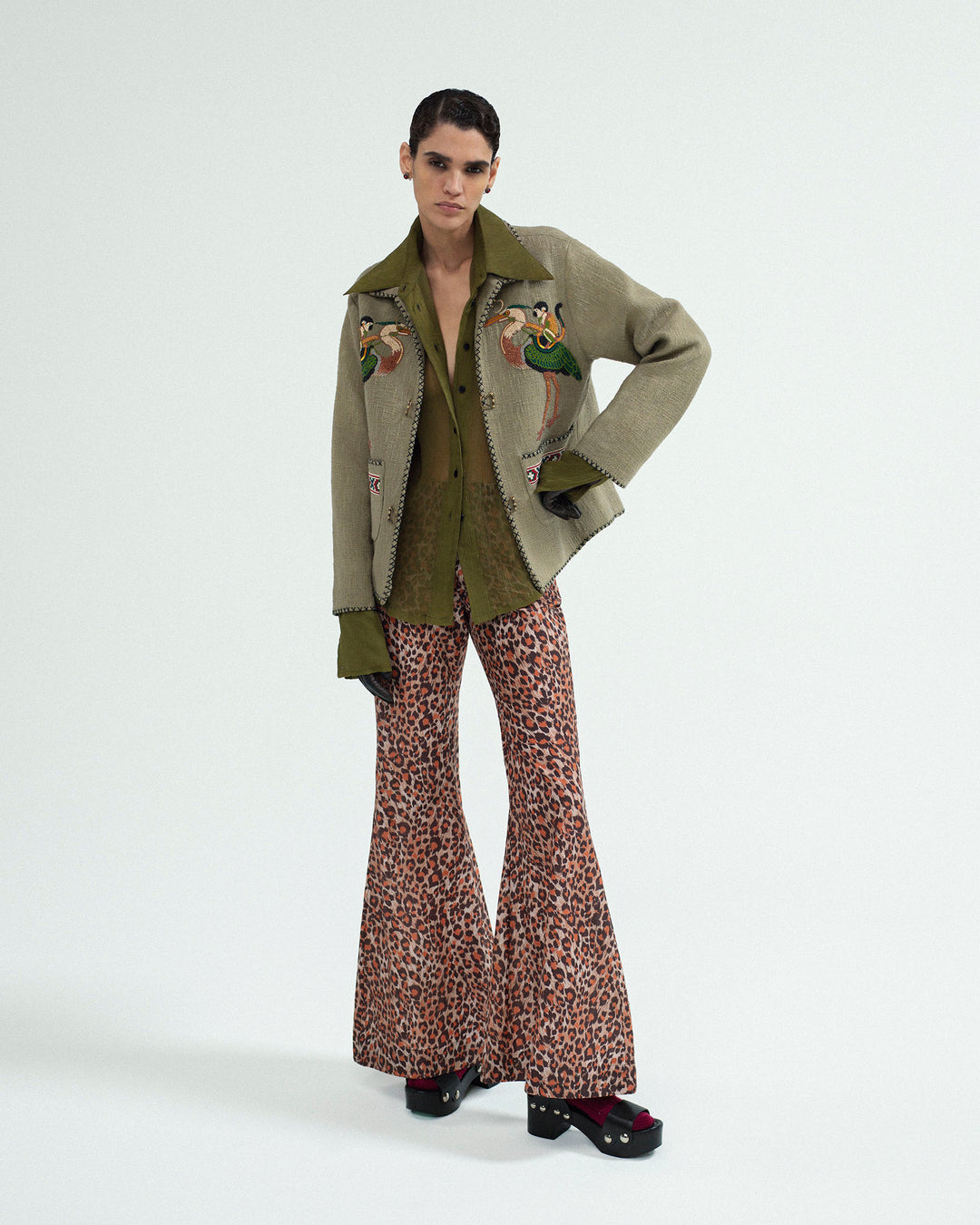 Wearing the Dana Olive Chiffon Shirt, an embroidered jacket, leopard-print flared pants, and black platform shoes, this person poses against a plain white background.