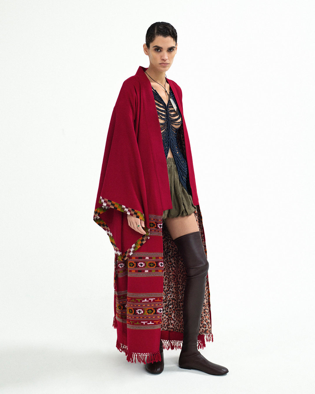 Person wearing the Grace Chili Tree Wool Coat—a red patterned cape reminiscent of a handwoven Kullu blanket—paired with a dark braided top, olive shorts, and thigh-high black boots stands against a plain white background.