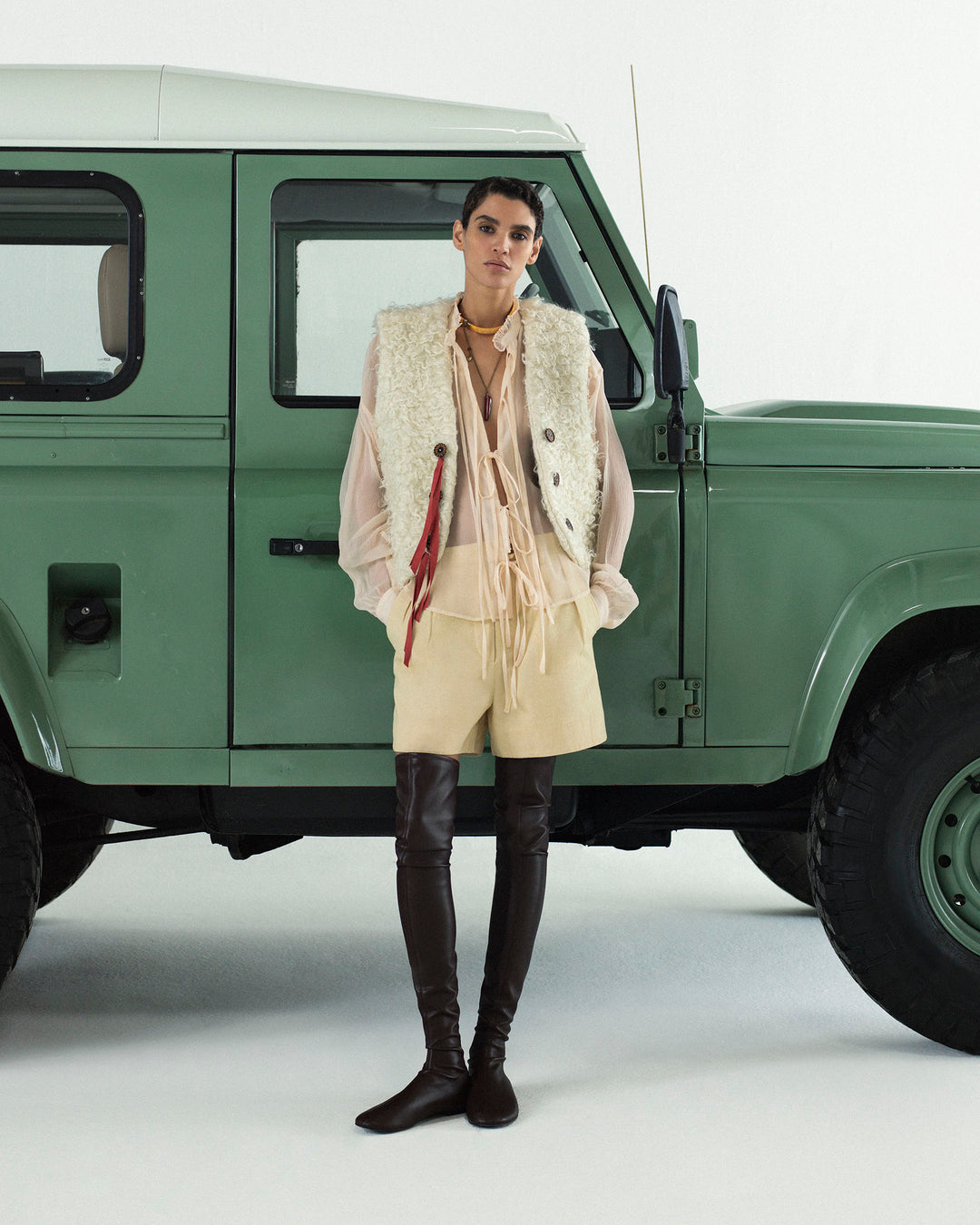 Person stands in front of a green SUV, wearing the Willa Prairie Tint Shirt—a cupro-chiffon sheer blouse with a subtle ruffled collar—layered under a cream vest, paired with beige shorts and thigh-high brown boots, hands tucked into their pockets.
