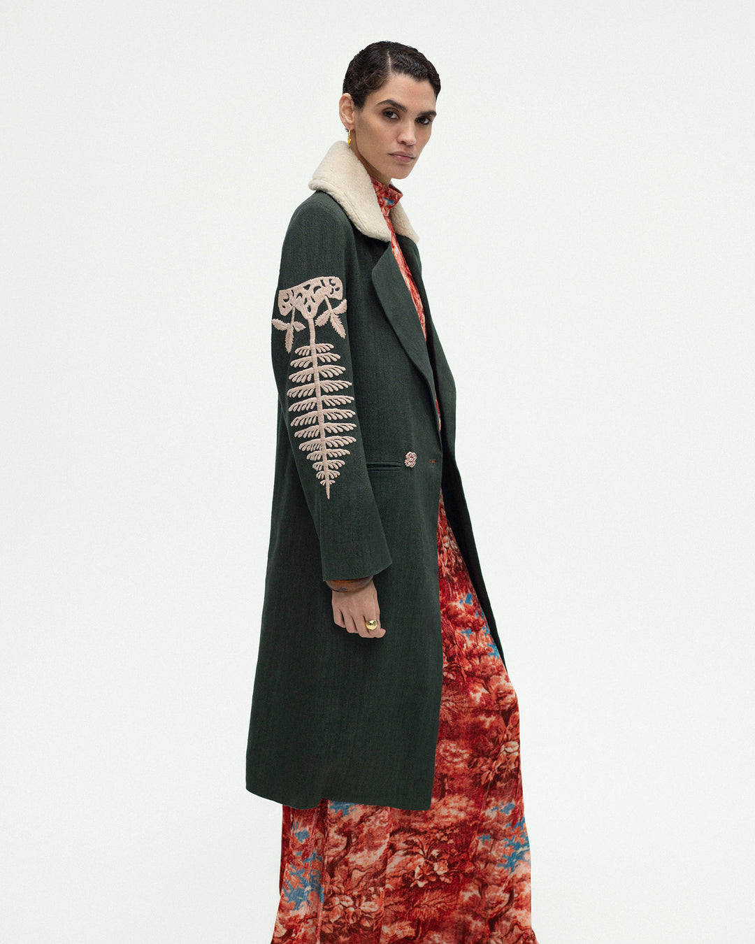 The Petunia Grassland Fern Coat, a long green wool-linen topcoat featuring a hand-embroidered white fern design and an Italian wool fleece collar, is layered over a red patterned dress and worn by a model standing against a plain white background.
