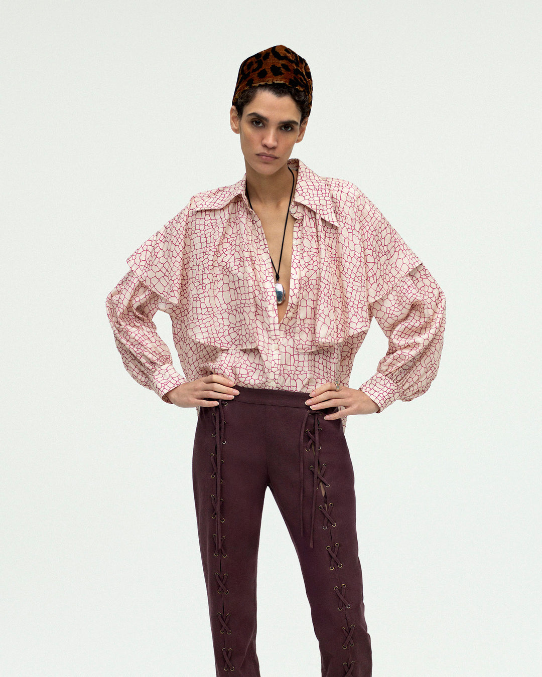 A person stands with hands on hips, wearing the Eleanor Crocodile Silk Shirt, dark lace-up pants, a necklace, and a leopard print hat against a plain light background.