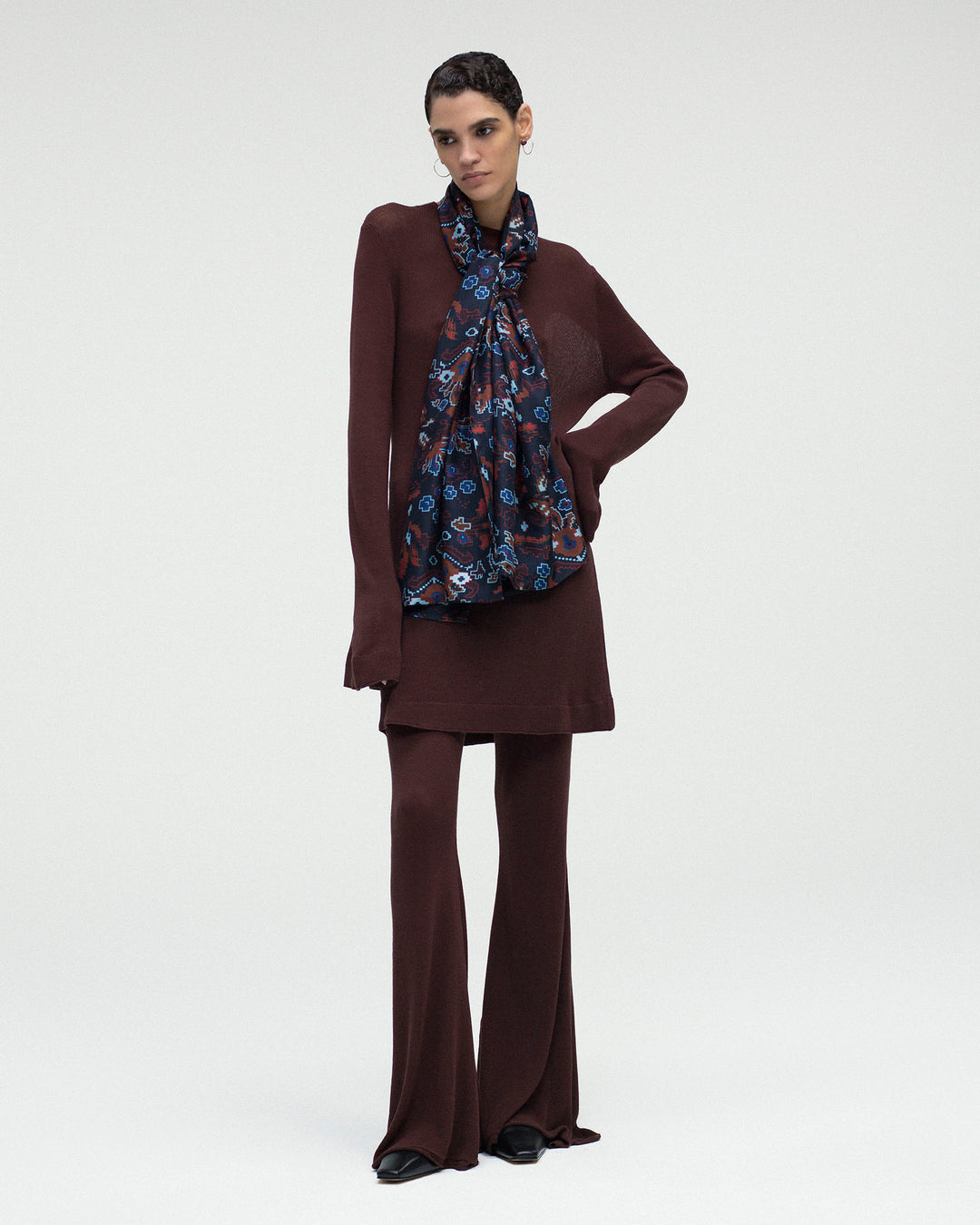 A model wearing a Romy Mahogany Knit Dress made of dark brown bamboo viscose with a long-sleeved tunic and matching flared trousers, accessorized with a dark blue patterned scarf.
