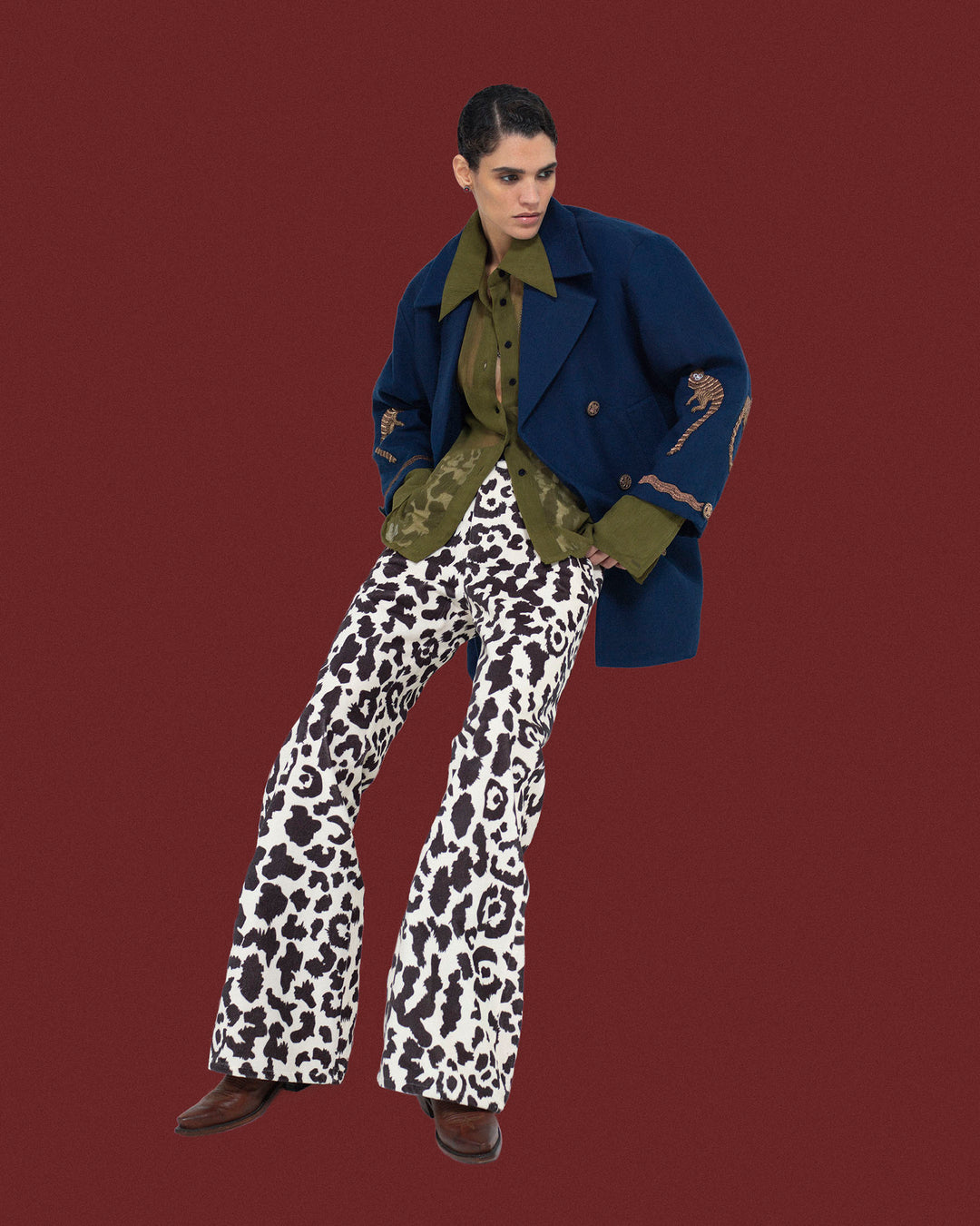 Wearing the Rivoli Midnight Mischief Coat, a person stands against a solid maroon background, paired with a green shirt and black-and-white animal print flared pants.