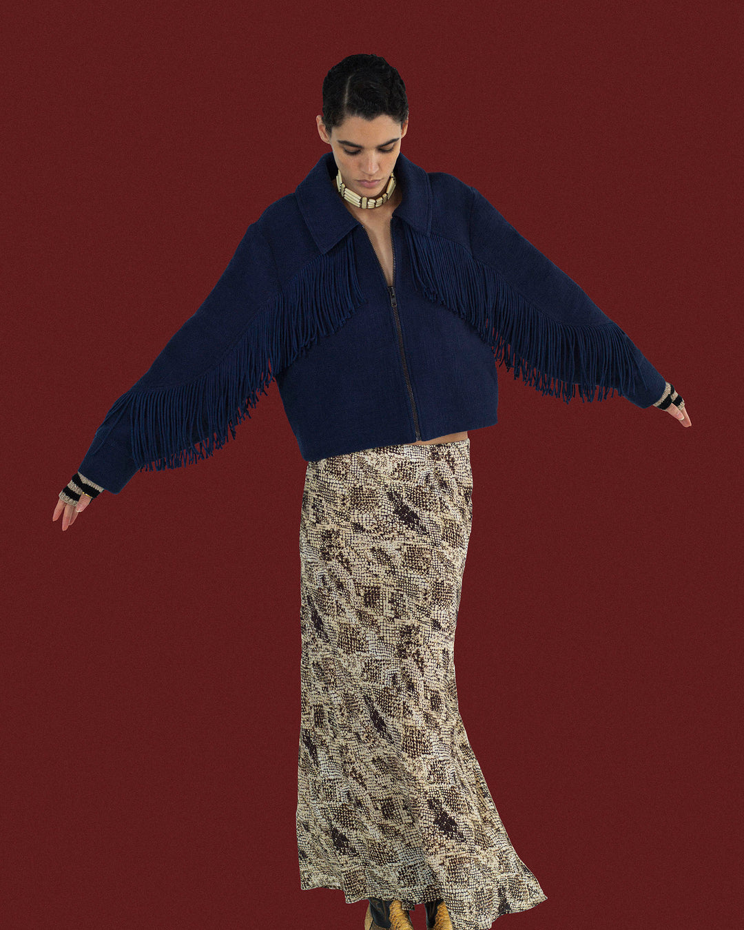 Wearing the Court Indigo Night Jacket, a person with arms outstretched stands against a solid maroon background, pairing the navy cropped zip-up with Western fringing and a long snake-print skirt.