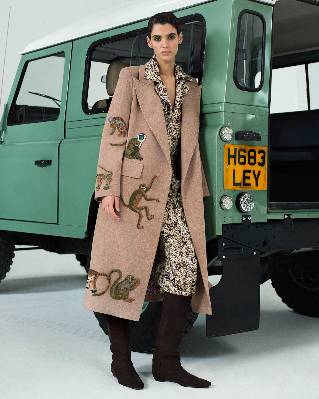 A person wearing the Duke Barrel of Monkeys Coat—a long tan wool herringbone topcoat with hand-beaded zoological motif—paired with brown boots stands in front of a vintage green vehicle with license plate H683 LEY.