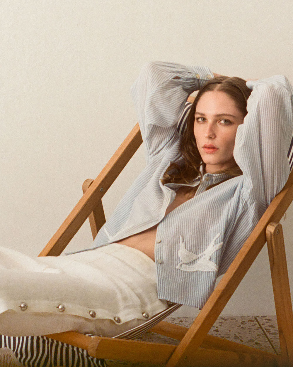 Wearing the Holland Pearl Linen Pant and a striped shirt, a person reclines on a wooden deck chair, gazing at the camera with a relaxed expression.