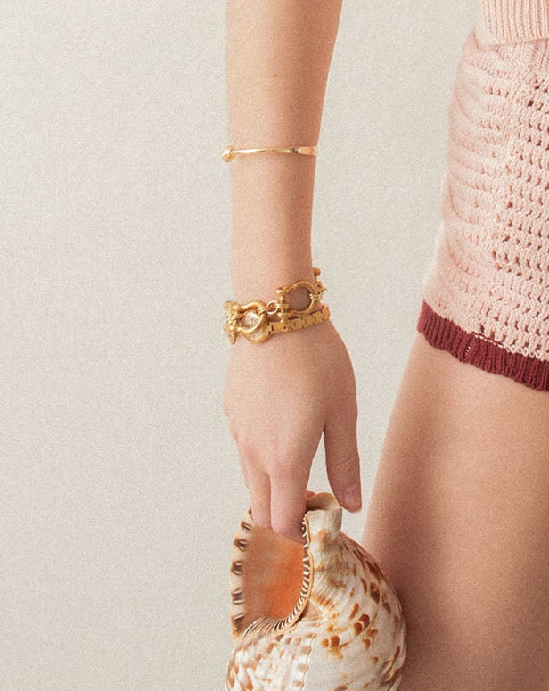Hand wearing a gold bracelet holding a seashell against a neutral background