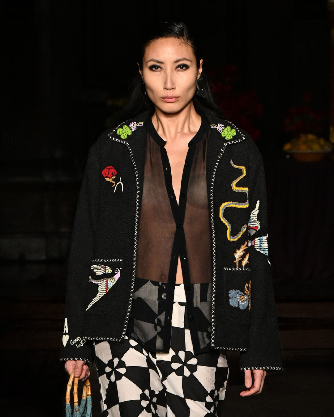 A model walks the runway wearing the Francis Black Tattoo Jacket over a sheer black shirt, black-and-white patterned pants, and holding a handbag.