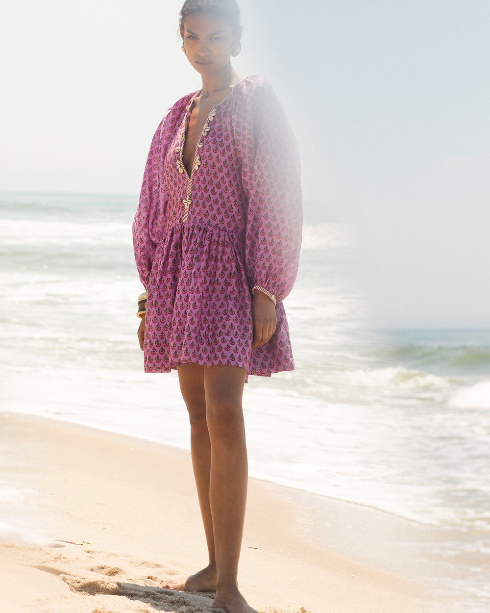 A person stands barefoot on a sandy beach, wearing the Sula Amethyst Dress, which features long sleeves embellished with cowrie shell accents. The ocean waves are visible in the background under a clear sky.