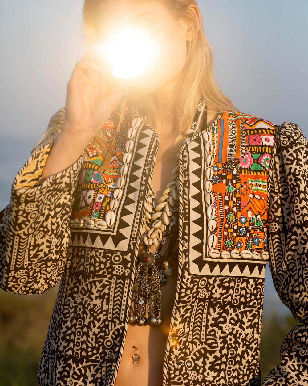 A person is outdoors, wearing the Hema Onyx Jacket, crafted with hand-printed cotton and adorned with embroidered trim. The intricate designs of the jacket feature vibrant colors and beadwork. The sunlight obscures the person's face, creating a glowing effect as it shines directly into the camera.