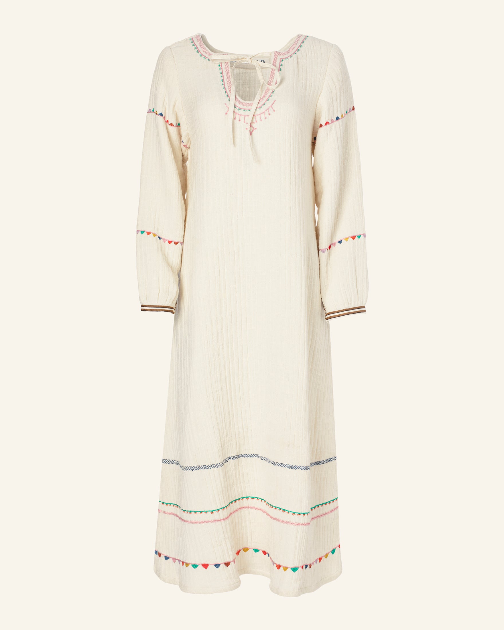 The Tali Embroidered Tunic Dress is a white, long-sleeve piece made from cotton gauze with a relaxed fit. It showcases vibrant hand-embroidered patterns around the neckline, sleeves, and hem, accented by a simple tie detail at the neck.