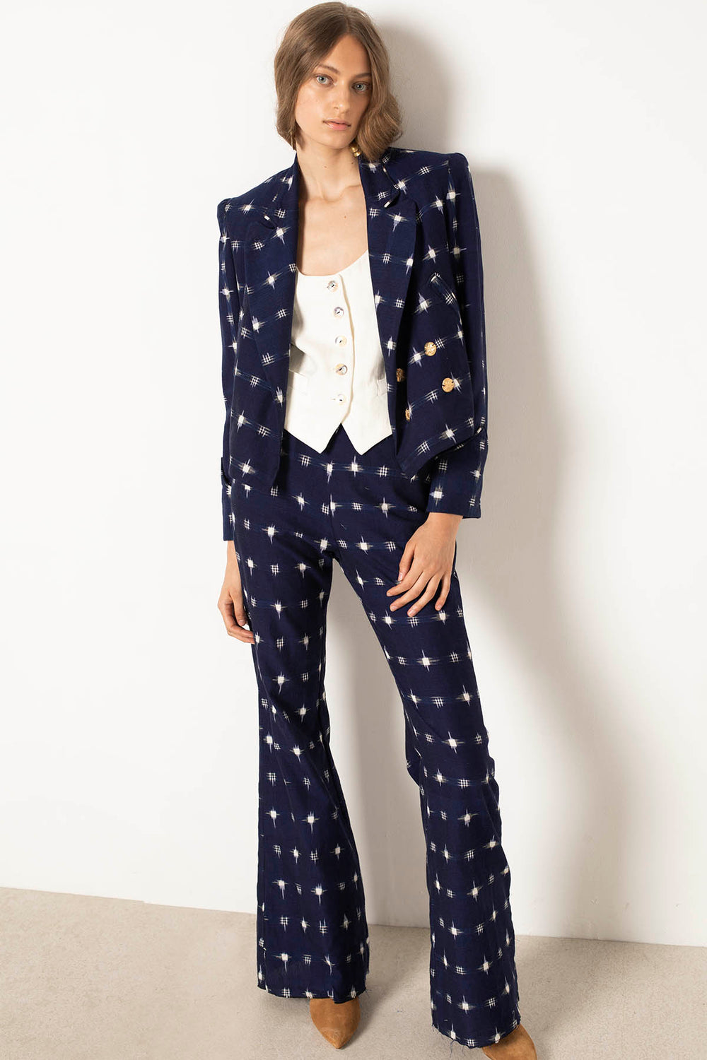 Against a white wall, a person with light brown hair stands in a stylish ensemble featuring the Charles Navy Ikat Spot Pant. The dark blue pantsuit showcases flared legs and an Indian ikat-inspired design. They sport a white buttoned vest and tan shoes, casually resting one hand in their pocket while maintaining a relaxed expression.