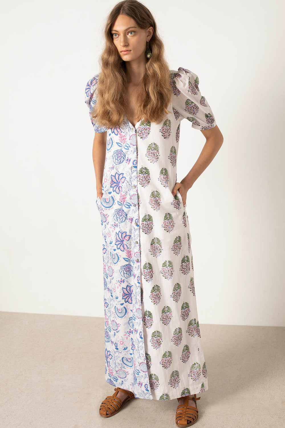 A woman with long hair is wearing the Fifty Fifty Bloom Dress, a floor-length, half-sleeve, A-line dress adorned with a floral pattern. One side showcases intricate blue flowers while the other displays purple floral motifs. The hand block-printed dress gracefully billows as she stands with her hands in her pockets.