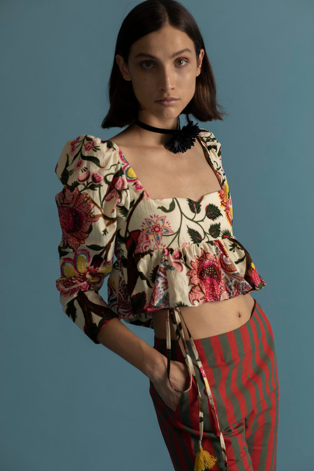 Against a blue backdrop, an individual wears the Lila Canary Peplum Top, featuring a floral pattern and puffed sleeves, paired with red striped pants. The outfit is accented by adjustable corset ties. They place their hands in their pockets and wear a black choker around their neck.