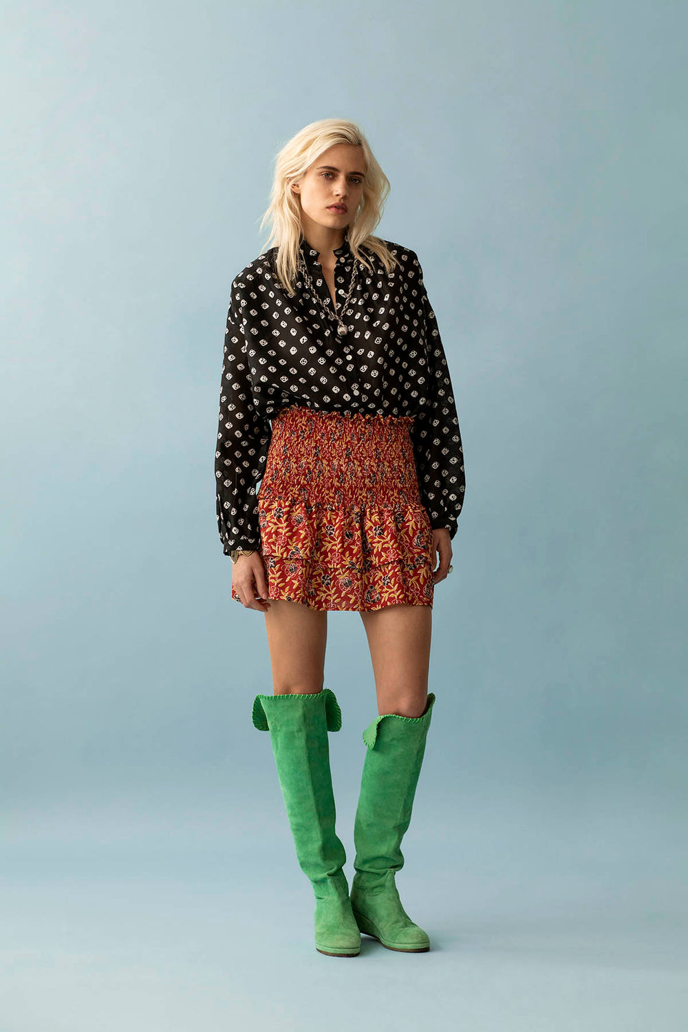 A person with long blond hair stands against a light blue background, showcasing unique beauty. They wear a black patterned blouse, the Maria Carnelian Ruffle Skirt—a red floral hand block printed cotton piece—and green knee-high boots.