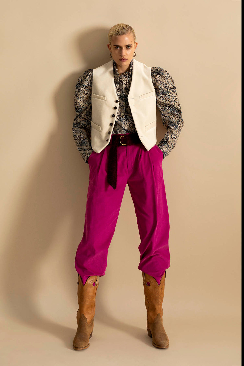 A person with slicked-back hair wears the Annabel Frost Shirt, a hand block printed floral cotton blouse featuring puffed sleeves and a ruffled collar. It's paired with a cream vest, bright pink pants, and cowboy boots. They stand confidently against a beige background with hands in pockets.