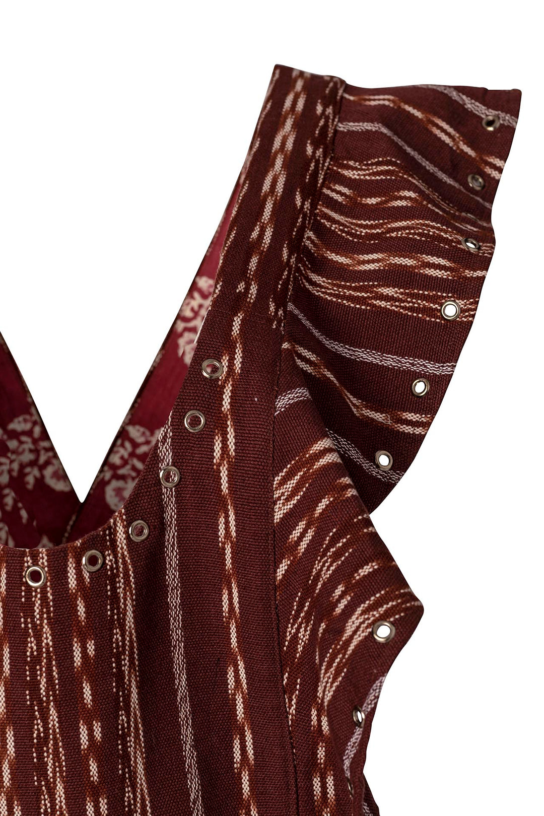 Close-up of the Indie Tobacco Ikat Overall, featuring a brown sleeveless design with white patterns and a ruffled edge, reminiscent of Guatemalan Ikat. The fabric showcases eyelet details that reveal a floral lining beneath, beautifully incorporating vertical stripes and wave-like patterns.