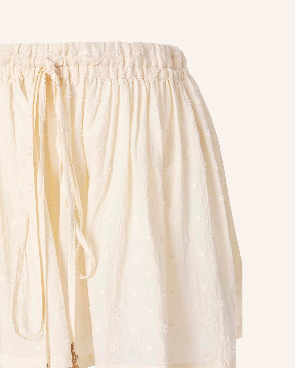 Introducing the Christy Powder Spot Short: this beige, textured skirt features an elasticated waistband with a drawstring for adjustable comfort. Crafted from Swiss Dot cotton, it boasts a subtle dotted pattern that provides a delicate touch. Its soft and lightweight design delivers a relaxed fit, making it an ideal choice for any casual occasion.