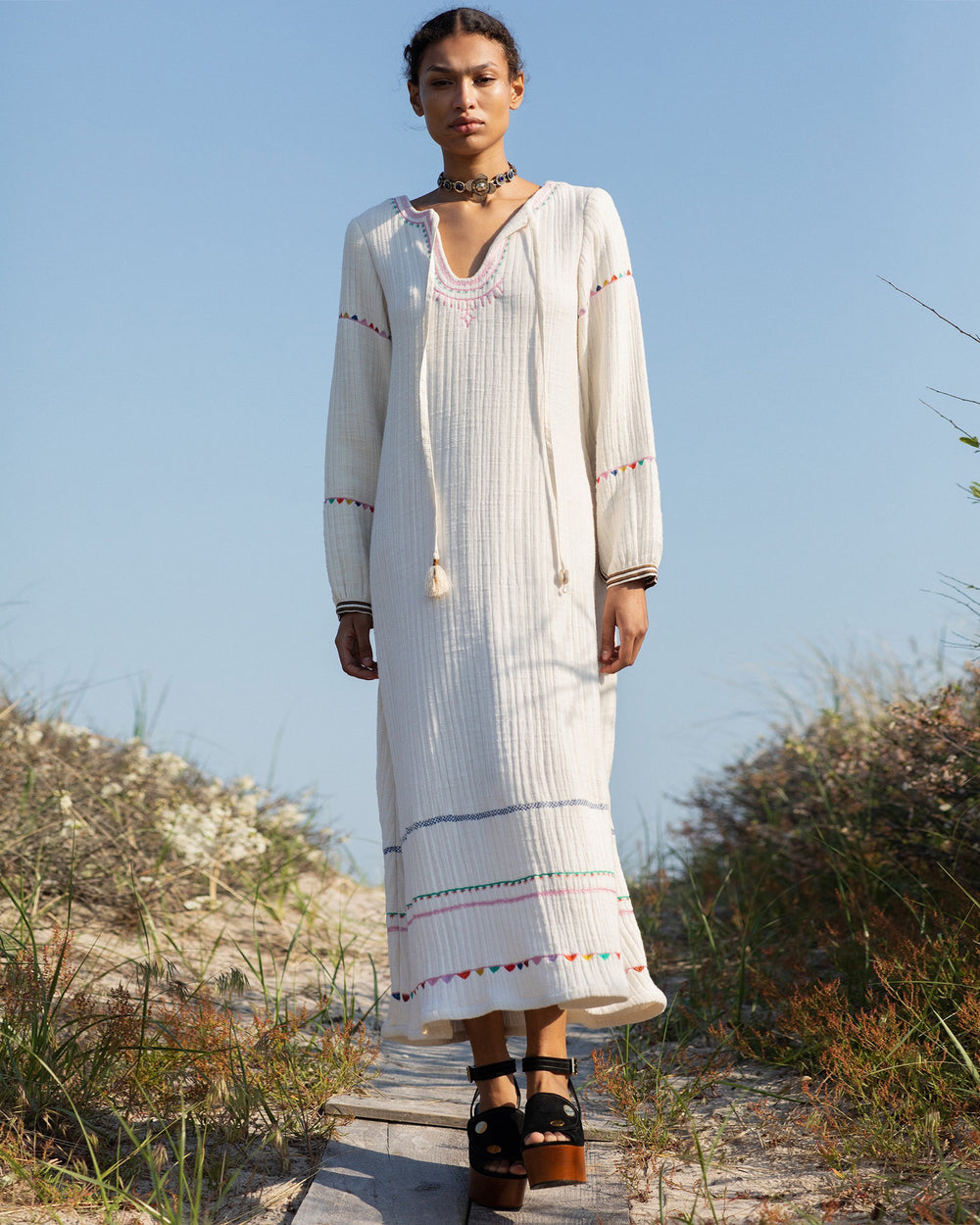 A person stands outdoors on a boardwalk wearing the Tali Embroidered Tunic Dress, made of cream-colored cotton gauze with colorful hand embroidery. They are also wearing platform sandals and a choker necklace, with a clear blue sky and coastal vegetation in the background.