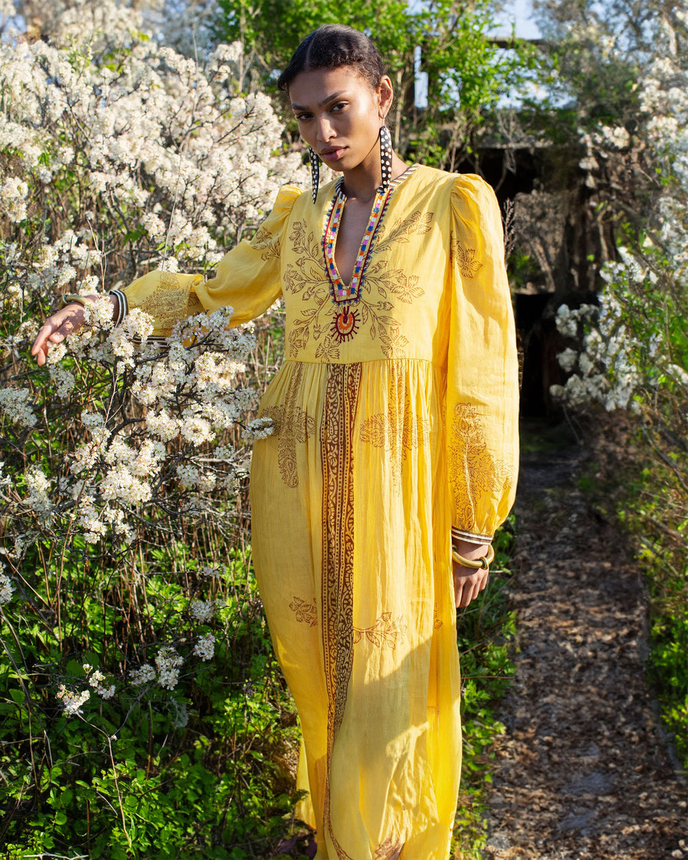 A person wearing the Winifred Amber Dust Dress, a long, hand-printed cotton yellow dress, stands in a garden surrounded by white flowering bushes. They have short, braided hair and are accessorized with jewelry, including a beaded necklace. The setting is lush and green.