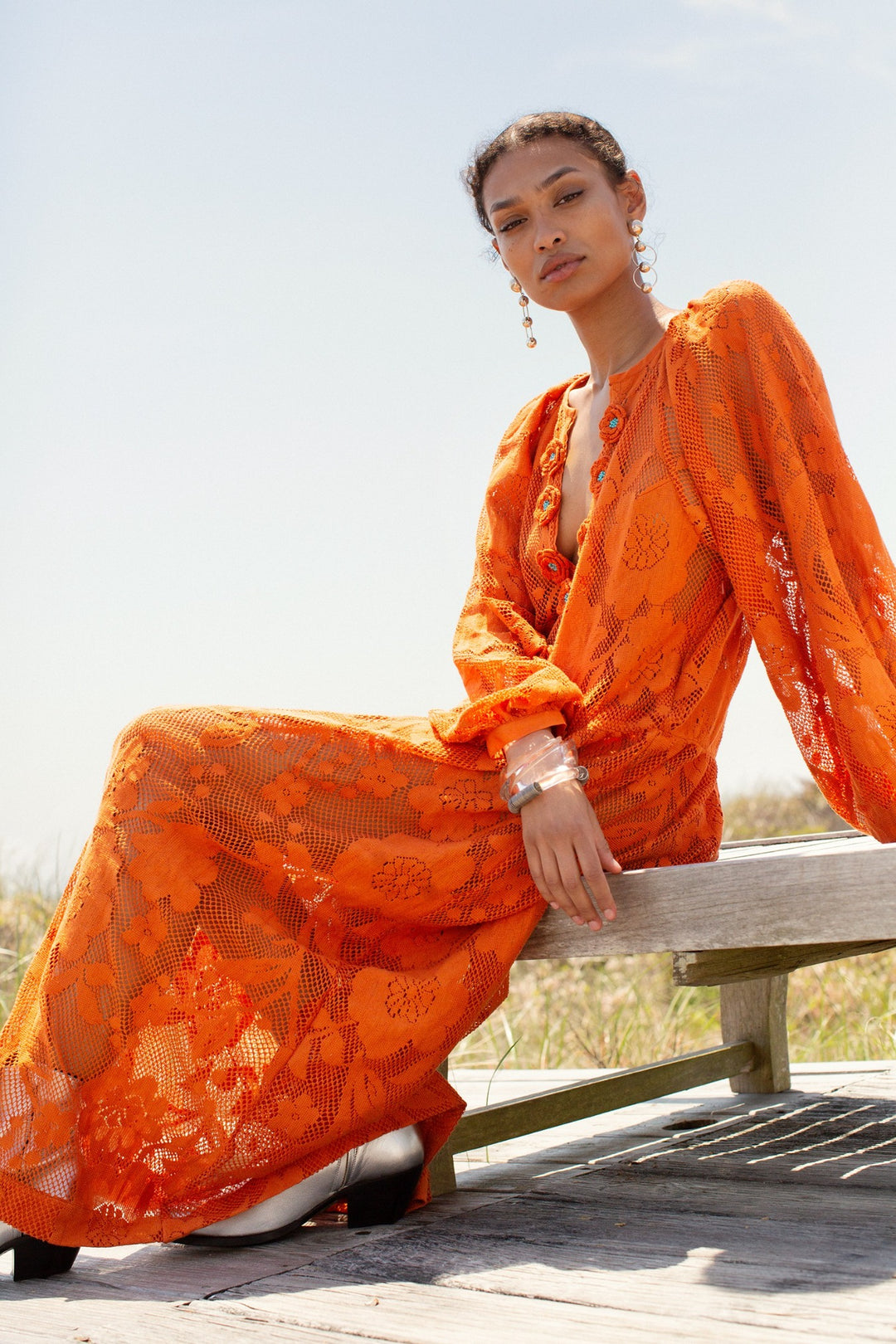 Wearing the Celeste Orangerie Lace Dress adorned with crochet flowers, a person sits on a wooden bench outdoors under a clear sky.