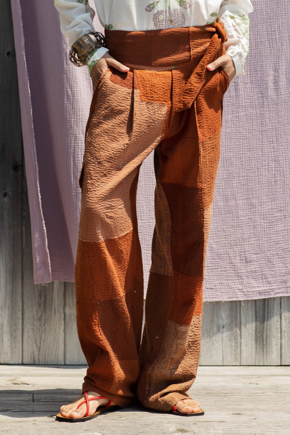 A person wearing the Georgia Earth Kantha Pant, featuring a wide and textured brown patchwork design, stands on a wooden floor. They also wear a white top with subtle floral embroidery, red sandals, and multiple bracelets. A colorful kantha quilt crafted from vintage saris hangs in the background.