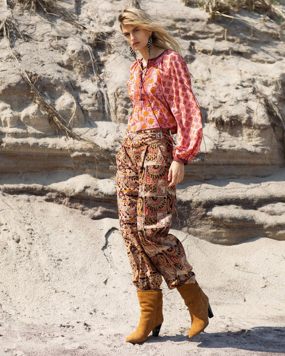 A person stands outdoors on sandy terrain, wearing a colorful outfit with a red patterned blouse and the Georgia Sunset Pant in hand-woven cotton. They have long, wavy hair and tan boots, looking off to the side.