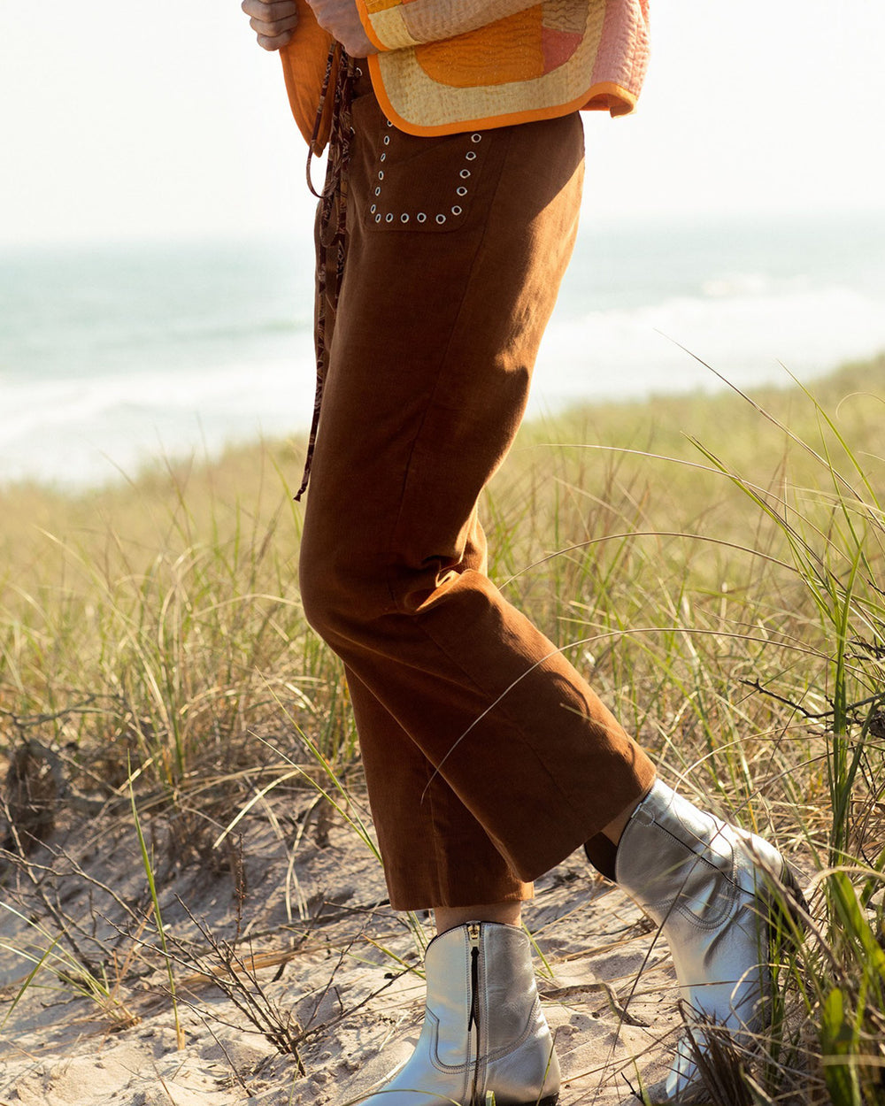 A person stands on a sandy beach with grass, wearing Jodhpur Tawny Corduroy Pants with grommet detailing, a plaid jacket, and shiny silver boots. The ocean and sky are visible in the background.