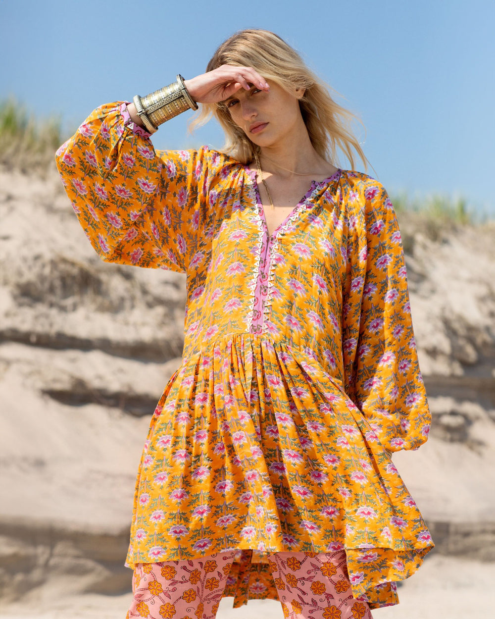 A person stands on a sandy beach in the Sula Turmeric Dress, a flowing yellow piece adorned with pink floral patterns, paired elegantly with hand-printed silk cotton pants. Their long blonde hair catches the breeze, while Mother of Pearl accents shimmer subtly from their bracelets as they shield their eyes from the clear sky.