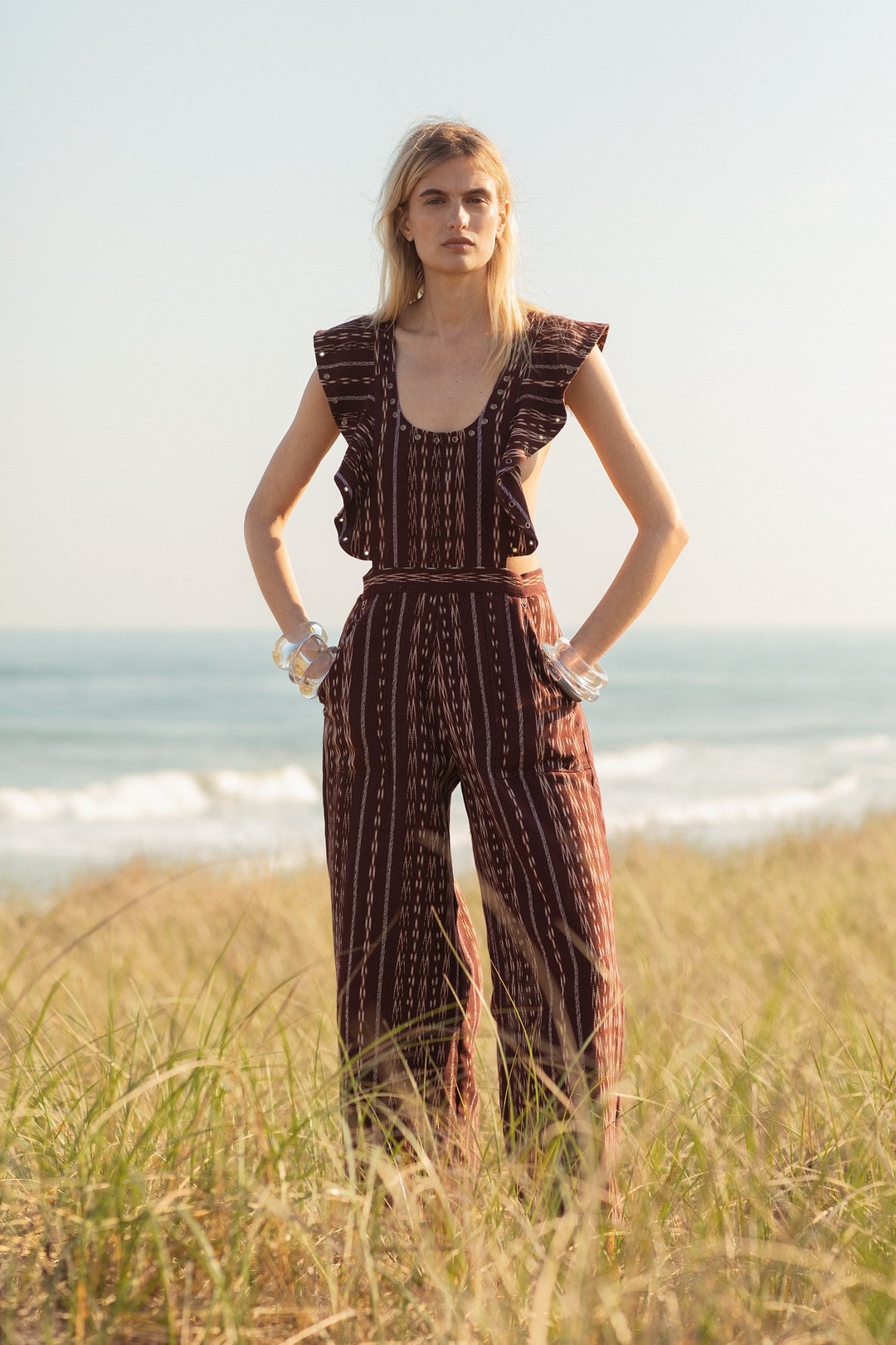 A person stands confidently on a beach with tall grass, wearing the Indie Tobacco Ikat Overall, showcasing ruffled sleeves and a design reminiscent of Guatemalan Ikat. The ocean is visible in the background under a clear sky.