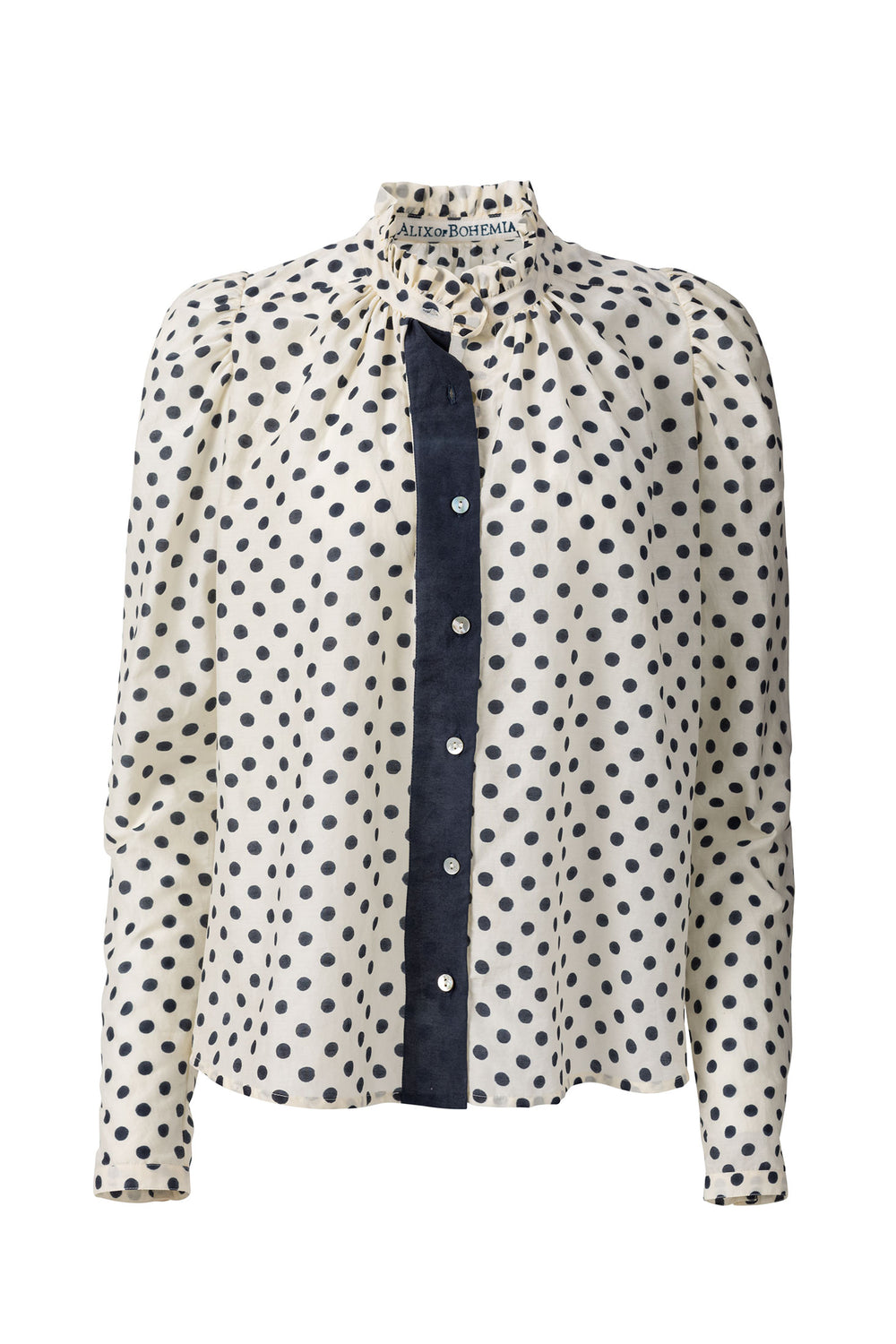 The Annabel Tuxedo Shirt is a white blouse adorned with black polka dots and features puffed sleeves. It showcases a high ruffled collar and a front button-down closure accented with a contrasting black panel.